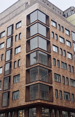 A modern multi-story building with a distinctive facade made of wooden shingles. The architecture features large glass windows and balconies creating a geometric pattern. Various levels of the building are visible, showcasing a consistent design with repeated window arrangements.