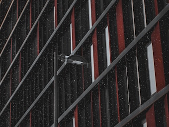 A modern facade with a grid of vertical and diagonal lines, featuring a predominantly dark surface with red and gray accents. A streetlight or security camera protrudes from the pattern, creating a focal point. Raindrops or speckles add texture across the surface.