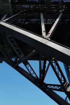 A close-up view of the understructure of a large metal bridge, showcasing intricate girders and beams. The geometric patterns formed by the intersecting steel elements create a sense of industrial strength and complexity. The sky in the background is clear and blue, providing contrast to the metallic texture.