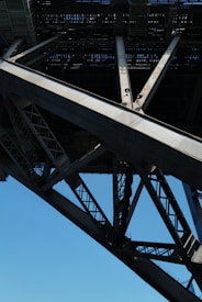 A close-up view of the understructure of a large metal bridge, showcasing intricate girders and beams. The geometric patterns formed by the intersecting steel elements create a sense of industrial strength and complexity. The sky in the background is clear and blue, providing contrast to the metallic texture.