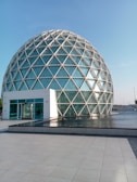 A geodesic dome with a large glass facade surrounded by a shallow pool of water, reflecting the structure. The dome is made up of triangular glass panels connected by metal frames. The foreground features a tiled courtyard leading to the water's edge.