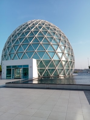 A geodesic dome with a large glass facade surrounded by a shallow pool of water, reflecting the structure. The dome is made up of triangular glass panels connected by metal frames. The foreground features a tiled courtyard leading to the water's edge.