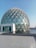 A geodesic dome with a large glass facade surrounded by a shallow pool of water, reflecting the structure. The dome is made up of triangular glass panels connected by metal frames. The foreground features a tiled courtyard leading to the water's edge.