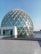 A geodesic dome with a large glass facade surrounded by a shallow pool of water, reflecting the structure. The dome is made up of triangular glass panels connected by metal frames. The foreground features a tiled courtyard leading to the water's edge.