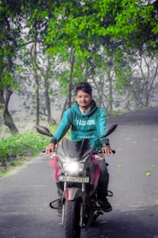 A person wearing a teal hoodie with the word 'FASHION' printed on it is riding a motorcycle down a road. The motorcycle's headlight is on. The road is bordered by lush green trees, and some fallen leaves are visible on the pavement. The surroundings suggest a scenic and serene environment.