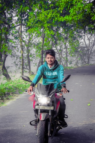A person wearing a teal hoodie with the word 'FASHION' printed on it is riding a motorcycle down a road. The motorcycle's headlight is on. The road is bordered by lush green trees, and some fallen leaves are visible on the pavement. The surroundings suggest a scenic and serene environment.