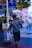 An older man is walking on a city street, pushing a cart filled with colorful toys and holding some balloons featuring cartoon characters. The scene takes place in an urban setting with trees, streetlights, and signs visible in the background.