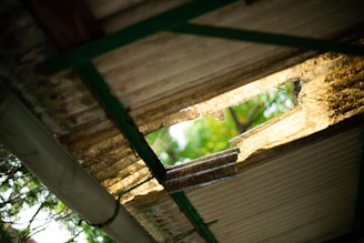 A close-up view of a corrugated metal roof with a large, jagged hole, allowing sunlight to filter through. The lighting casts shadows and highlights the rust and wear on the metal. In the background, blurred green foliage is visible, adding a natural element.