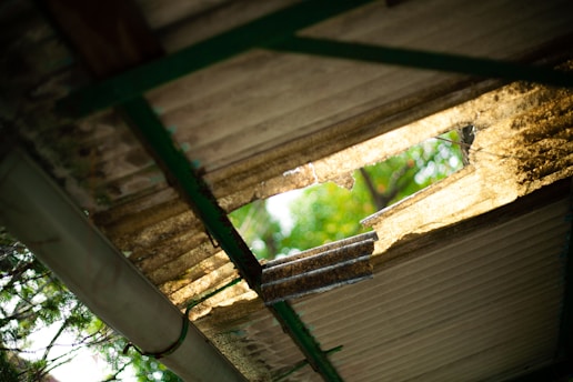 A close-up view of a corrugated metal roof with a large, jagged hole, allowing sunlight to filter through. The lighting casts shadows and highlights the rust and wear on the metal. In the background, blurred green foliage is visible, adding a natural element.
