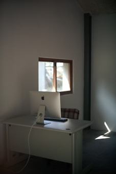 A minimalist workspace featuring a modern computer on a plain white desk, positioned near a small window with natural light illuminating the room. The space is simple, with a single chair, and the walls are bare, emphasizing the clean aesthetic.