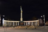 A grand monument featuring a tall column topped with a statue illuminated against a dark night sky. The base of the monument is surrounded by an arcade of columns with lights casting a warm glow. Sparse streetlights and trees are visible, along with a lone figure sitting nearby.