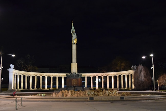 A grand monument featuring a tall column topped with a statue illuminated against a dark night sky. The base of the monument is surrounded by an arcade of columns with lights casting a warm glow. Sparse streetlights and trees are visible, along with a lone figure sitting nearby.