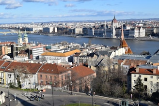 A cityscape featuring the iconic Parliament building along a riverbank, with a prominent gothic-style church and other historical architecture in the foreground. The river divides the city, and there are signs of modern urban life with people walking along the streets. The sky is partly cloudy, and the overall view is expansive showcasing both old and new buildings.