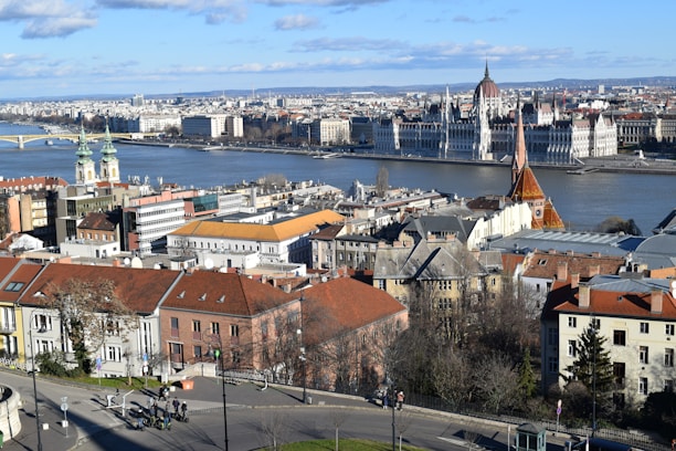 A cityscape featuring the iconic Parliament building along a riverbank, with a prominent gothic-style church and other historical architecture in the foreground. The river divides the city, and there are signs of modern urban life with people walking along the streets. The sky is partly cloudy, and the overall view is expansive showcasing both old and new buildings.