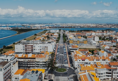 Aerial view of a coastal town with a prominent river or canal dividing two sections of land. The town features a mix of residential buildings with orange-tiled roofs, and streets lined with parked cars. In the background, industrial areas and distant landmasses are visible across the water. The sky is partly cloudy, adding to the scenic landscape.