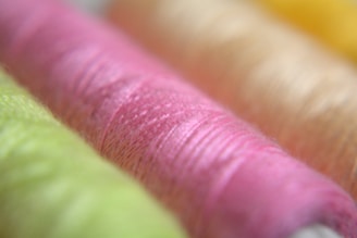 Colorful spools of thread in soft focus, displaying vibrant shades of green, pink, beige, and yellow. The threads are arranged closely, emphasizing the texture and fine details of the fibers.