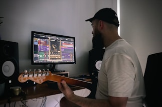 A person wearing a cap and a white shirt is seated at a desk, holding a guitar and looking at a computer screen displaying music production software. There are two speakers on either side of the monitor on the desk.
