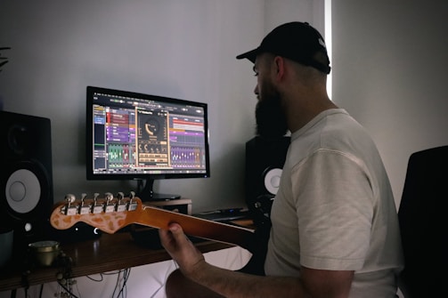 A person wearing a cap and a white shirt is seated at a desk, holding a guitar and looking at a computer screen displaying music production software. There are two speakers on either side of the monitor on the desk.