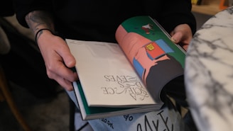 A person is holding a book open, featuring colorful illustrations on the right page and text on the left. The person's hands are visible, with tattoos on one wrist and chipped nail polish. A marble tabletop and part of the person's pants, which appear to have text or graphics, are also seen in the image.
