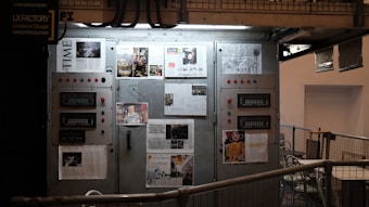 An industrial setting featuring a large metallic control panel adorned with various newspaper clippings and articles. Several panels with buttons and indicators are visible, surrounded by metal railings and pipes. The words 'LX FACTORY' are visible on a sign above, suggesting this is a creative or cultural space.