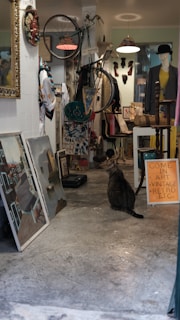An eclectic shop interior filled with various items including a bicycle mounted on the wall, several pieces of artwork leaning against a wall, a mannequin wearing clothing, and a tabby cat sitting on the floor. The shop has a vintage vibe, with a sign inviting people to come in for art and retro items. The lighting is warm, and there are various decorative elements scattered throughout.