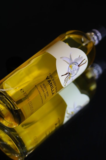 A bottle of body mist labeled 'Vanilla' with a floral design on the label. The bottle is transparent with a yellow-tinted liquid inside and is positioned on a reflective black surface.