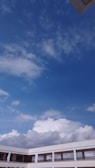 A large expanse of blue sky scattered with fluffy white clouds is visible above the rooftops of a modern building. The building has a white exterior with multiple rows of windows and railings, suggesting a multi-story structure, possibly a school or office.