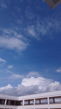 A large expanse of blue sky scattered with fluffy white clouds is visible above the rooftops of a modern building. The building has a white exterior with multiple rows of windows and railings, suggesting a multi-story structure, possibly a school or office.