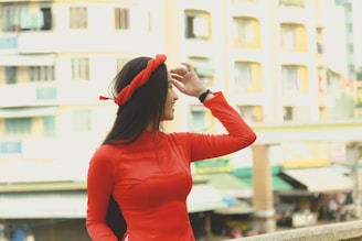 A woman in a vibrant red outfit and headband appears to be smiling while looking into the distance with her hand shielding her eyes. She stands against a backdrop of tall buildings and urban scenery.