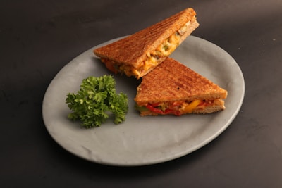 Two grilled cheese sandwiches stuffed with vegetables, placed on a grayish-white plate with a garnish of fresh parsley on a dark background.