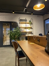 A modern office space features a wooden desk with a blue-cushioned chair and various objects such as a laptop bag. In the background, there is a white brick wall with a black metal and wood bookshelf adorned with books and green plants. A large potted plant sits near a glass door. Warm lighting is provided by a ceiling lamp.