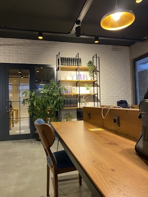A modern office space features a wooden desk with a blue-cushioned chair and various objects such as a laptop bag. In the background, there is a white brick wall with a black metal and wood bookshelf adorned with books and green plants. A large potted plant sits near a glass door. Warm lighting is provided by a ceiling lamp.