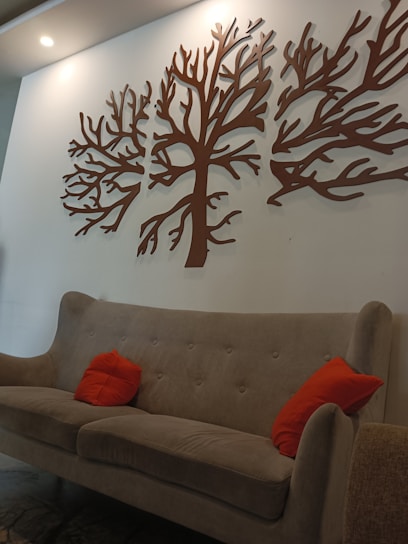A modern interior setting features a light brown upholstered sofa adorned with two bright orange cushions. Above the sofa, the wall showcases a decorative piece of art consisting of abstract tree branches crafted in brown material. The room is softly lit by overhead lighting, adding a warm ambiance.