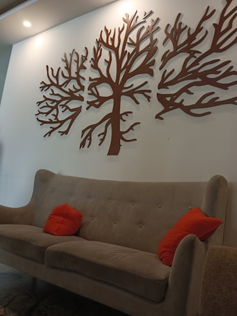 A modern interior setting features a light brown upholstered sofa adorned with two bright orange cushions. Above the sofa, the wall showcases a decorative piece of art consisting of abstract tree branches crafted in brown material. The room is softly lit by overhead lighting, adding a warm ambiance.
