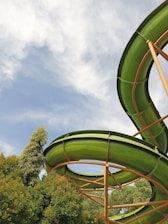 A vibrant green water slide curves against a backdrop of a blue sky with white clouds. Lush green trees surround the slide, creating a natural and inviting atmosphere.