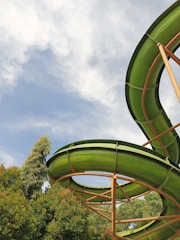 A vibrant green water slide curves against a backdrop of a blue sky with white clouds. Lush green trees surround the slide, creating a natural and inviting atmosphere.