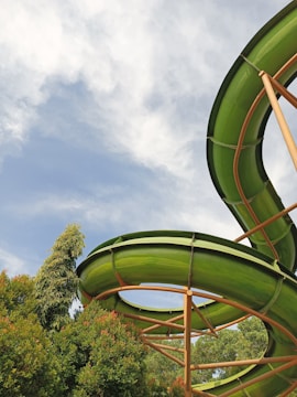 A vibrant green water slide curves against a backdrop of a blue sky with white clouds. Lush green trees surround the slide, creating a natural and inviting atmosphere.