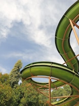 A vibrant green water slide curves against a backdrop of a blue sky with white clouds. Lush green trees surround the slide, creating a natural and inviting atmosphere.