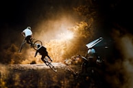Two cyclists are captured mid-action, performing stunts on a dirt trail surrounded by a forest. The scene is bathed in warm, golden sunlight, highlighting the energy and skill of the riders. A group of spectators and photographers can be seen watching from the side, with one person taking a photo. The setting conveys a sense of adventure and sport.