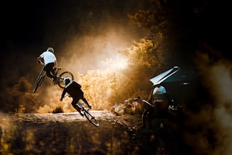 Two cyclists are captured mid-action, performing stunts on a dirt trail surrounded by a forest. The scene is bathed in warm, golden sunlight, highlighting the energy and skill of the riders. A group of spectators and photographers can be seen watching from the side, with one person taking a photo. The setting conveys a sense of adventure and sport.