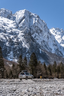 A rugged mountain landscape featuring towering snow-capped peaks under a clear blue sky. In the foreground, a white SUV is parked on a rocky terrain surrounded by pine trees. A person in yellow pants and a dark jacket stands nearby, seemingly taking a photograph.