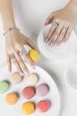 A pair of hands with manicured nails and adorned with intricate rings and a bracelet is seen interacting with colorful macarons. The macarons are arranged on white plates, showcasing a variety of pastel shades including yellow, pink, green, and peach.