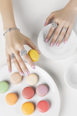 A pair of hands with manicured nails and adorned with intricate rings and a bracelet is seen interacting with colorful macarons. The macarons are arranged on white plates, showcasing a variety of pastel shades including yellow, pink, green, and peach.