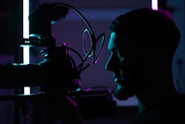 A silhouetted profile of a person holding a professional camera, illuminated by contrasting blue and purple lights, creating a dramatic and artistic atmosphere.