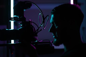 A silhouetted profile of a person holding a professional camera, illuminated by contrasting blue and purple lights, creating a dramatic and artistic atmosphere.