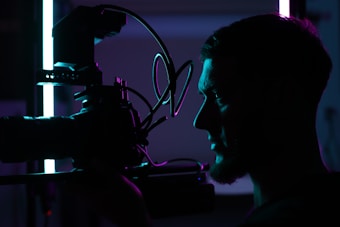 A silhouetted profile of a person holding a professional camera, illuminated by contrasting blue and purple lights, creating a dramatic and artistic atmosphere.