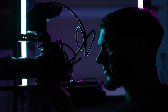 A silhouetted profile of a person holding a professional camera, illuminated by contrasting blue and purple lights, creating a dramatic and artistic atmosphere.