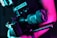 A professional camera held by a person is illuminated in vibrant neon pink and teal lighting. The camera has a large lens and appears to be mounted on a stabilizer or rig, indicating it's suitable for high-quality video or film production.
