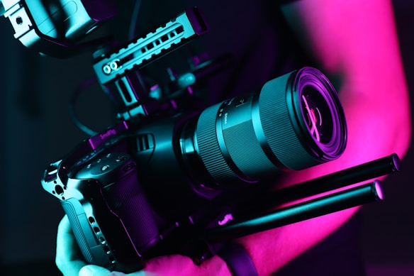 A professional camera held by a person is illuminated in vibrant neon pink and teal lighting. The camera has a large lens and appears to be mounted on a stabilizer or rig, indicating it's suitable for high-quality video or film production.