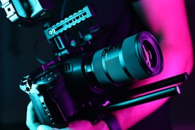 A professional camera held by a person is illuminated in vibrant neon pink and teal lighting. The camera has a large lens and appears to be mounted on a stabilizer or rig, indicating it's suitable for high-quality video or film production.
